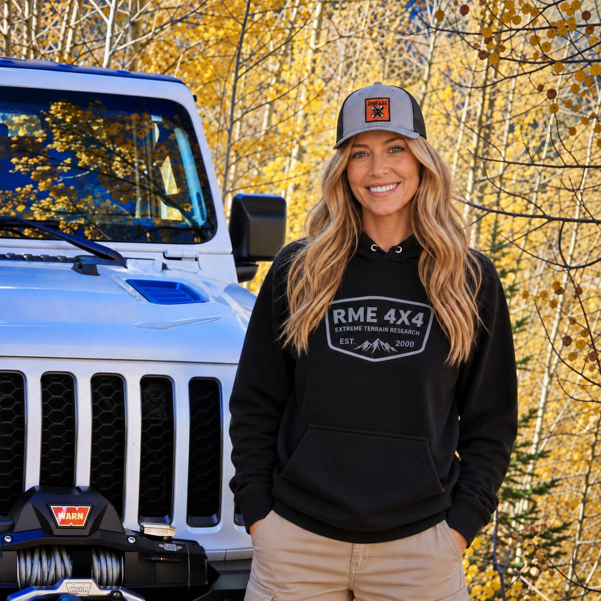 Woman in an RME 4X4 hoodie and U-joint hat next to a white Jeep in the aspens
